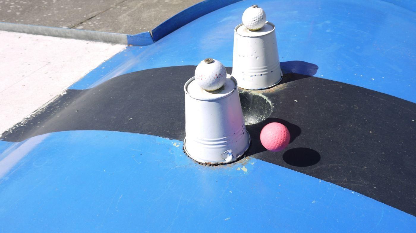 Blaue Minigolfbahn mit zwei weißen Bechern als Hindernis, daneben ein pinker Ball