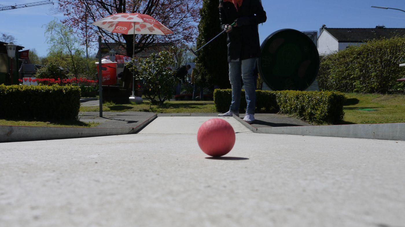 Pinker Minigolfball auf einer Bahn der Minigolf-Oase, im Hintergrund ein Spieler, Hecken und ein rot-weißer Sonnenschirm