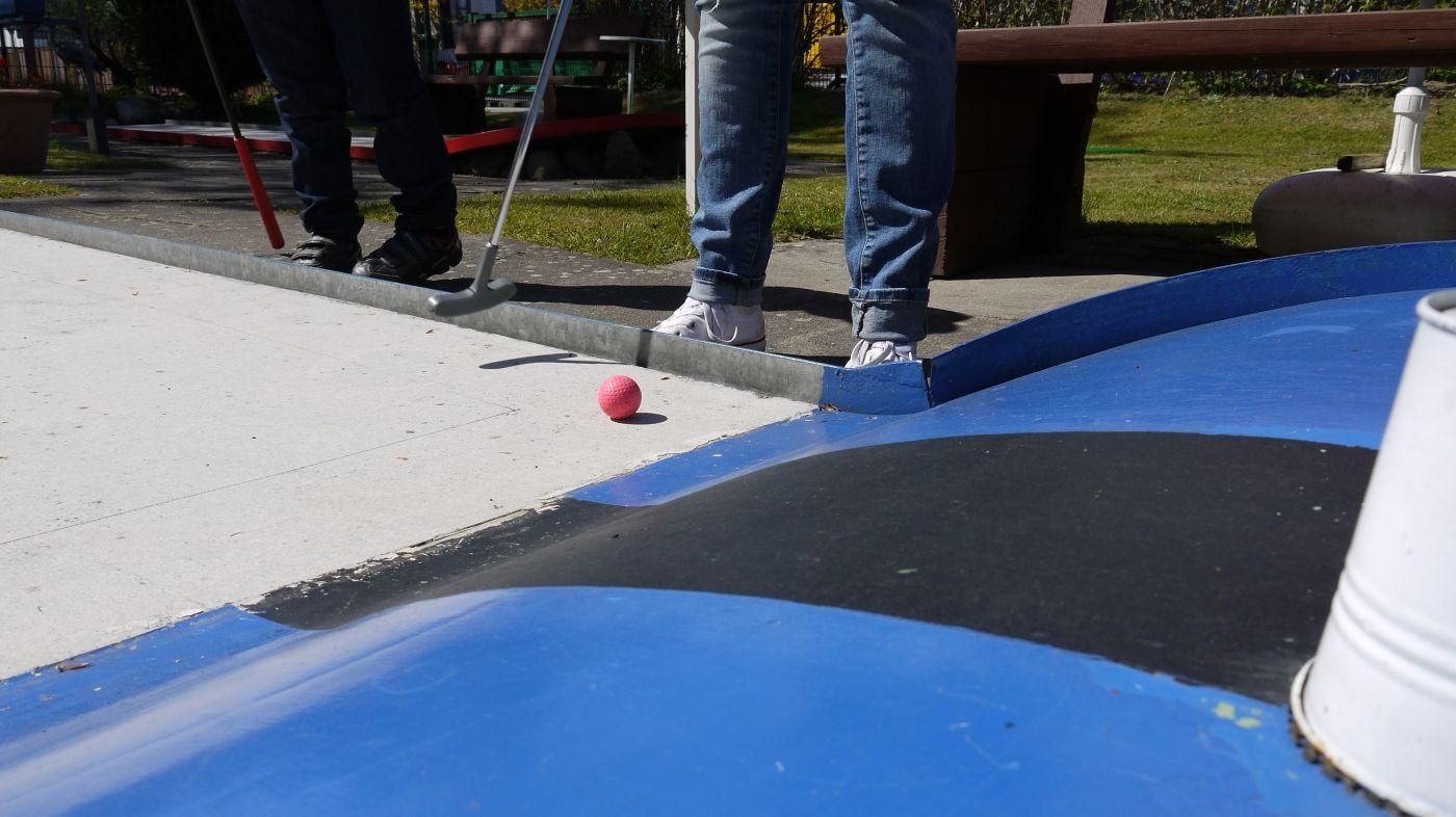 Pinker Ball kurz vor dem Loch, daneben die Füße der Spieler und eine blaue Bande