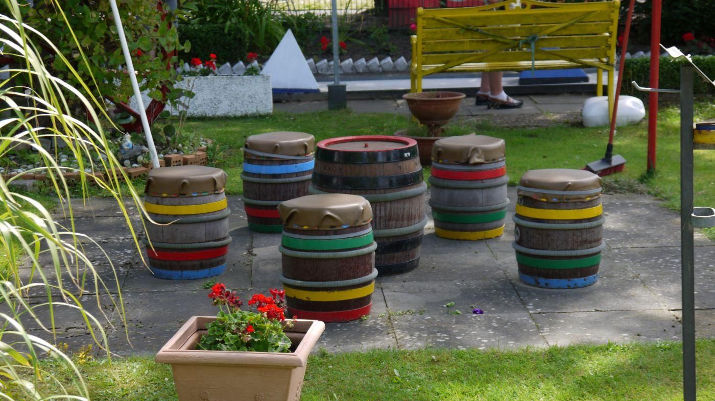 Bunt bemalte Fässer als Hindernis auf einer Minigolfbahn, davor ein Blumenkübel mit roten Geranien