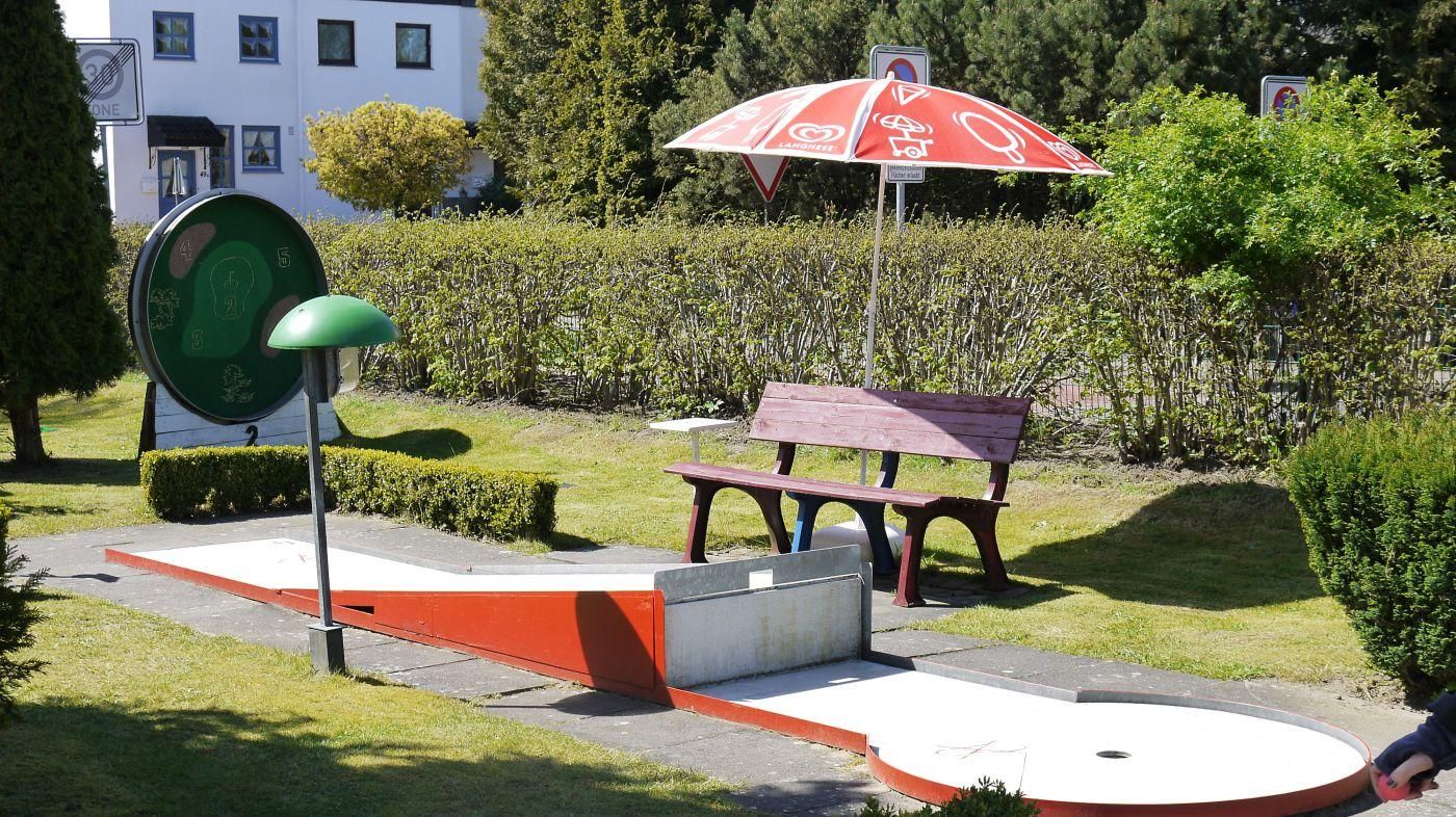 Minigolfbahn mit Hindernis, dahinter eine Bank und ein Sonnenschirm