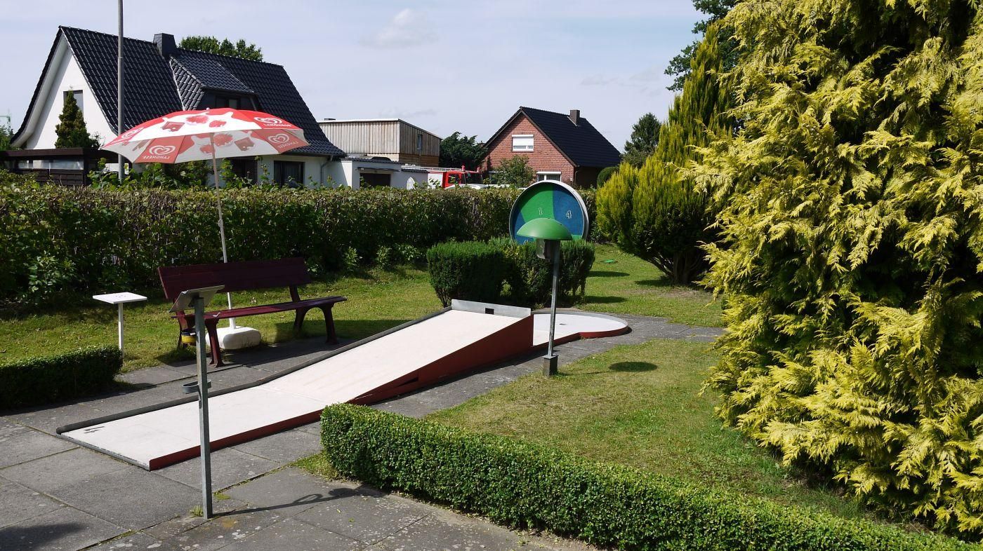Minigolfbahn mit Rampe zwischen Hecken und Rasen, im Hintergrund ein rot-weißer Sonnenschirm