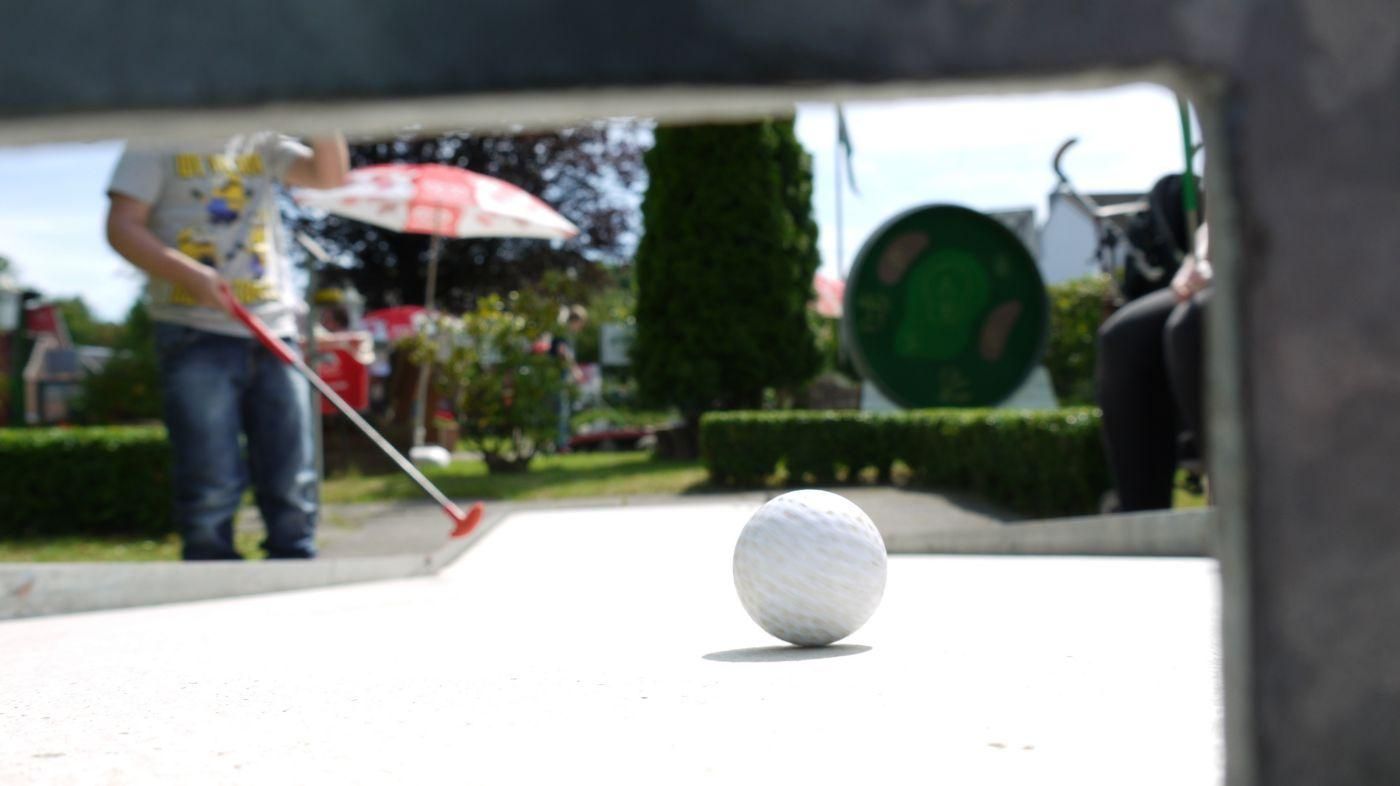 Weißer Minigolfball auf der Bahn, dahinter ein Kind beim Schlag
