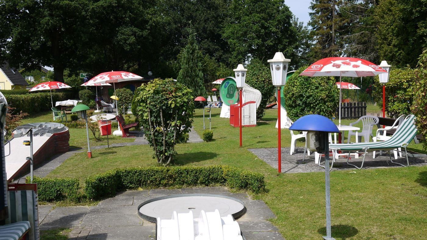 Blick über die Minigolfanlage mit Laternen, rot-weißen Sonnenschirmen und Bahnen zwischen Hecken